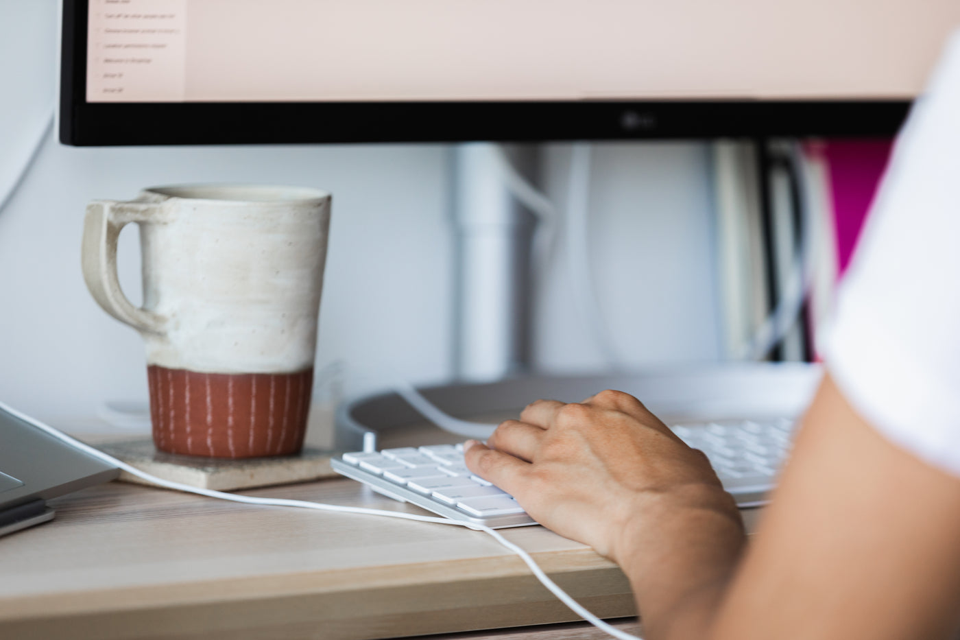 Lifestyle image of a close up of a person's hand on a keyboard. The bottom of a monitor is visible and there's a coffee cup on a coaster.